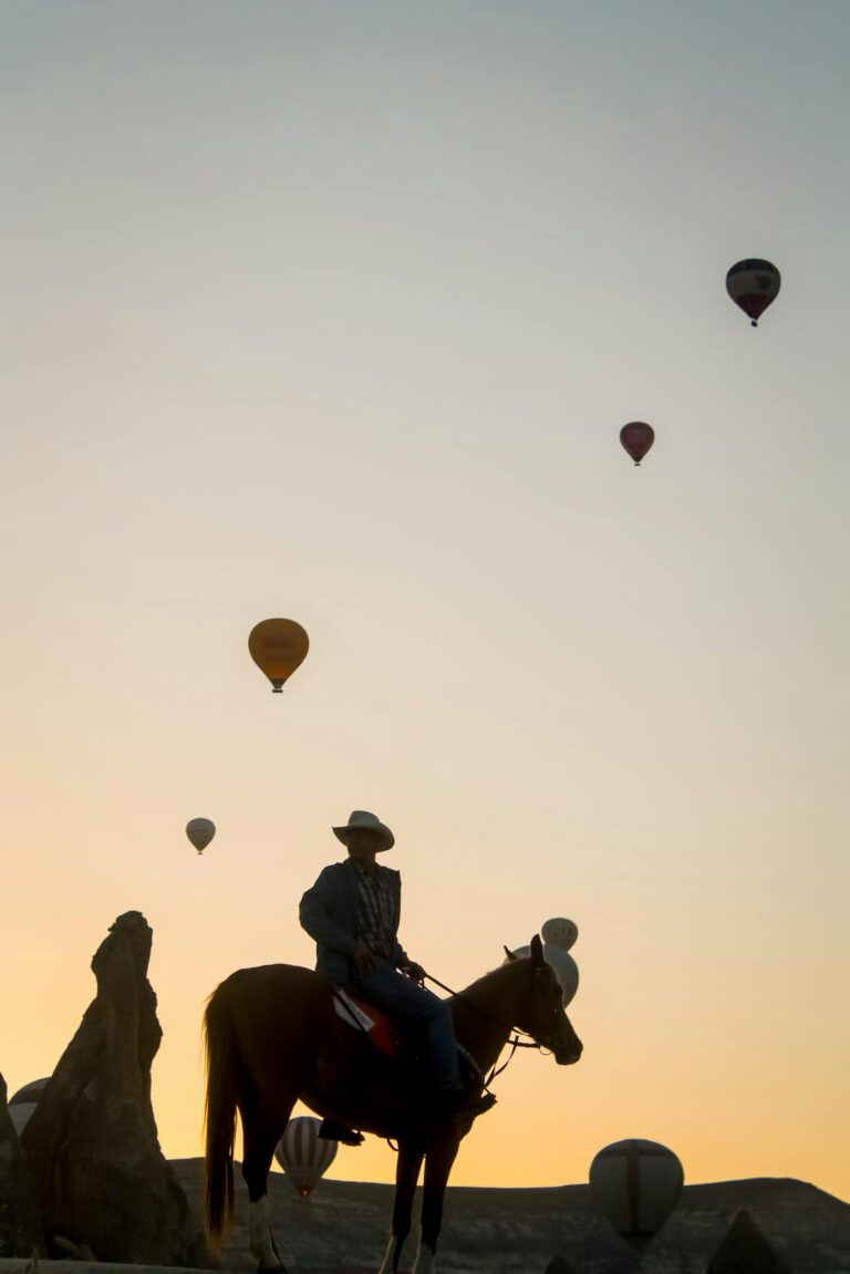 A silhouette of a horse rider at sunset with hot air balloons in the sky over Cappadocia, Türkiye.