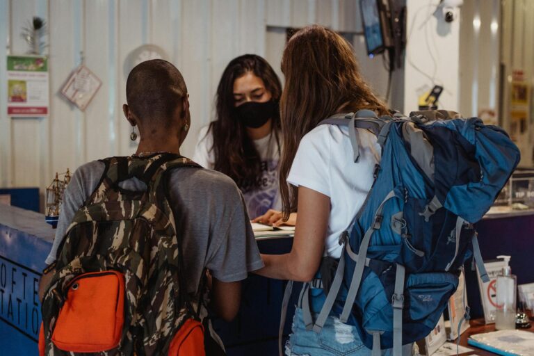 Two backpackers checking in at a hostel reception, wearing face masks.